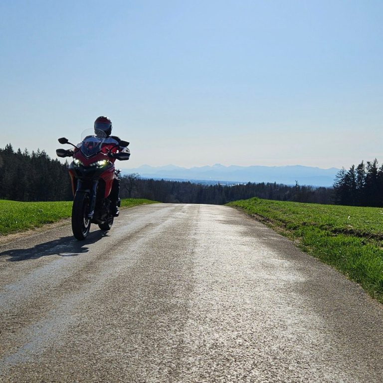 KurvenScout Ein Motorrad steht auf einer schmalen, sonnigen Straße mit Grünflächen und die Chiemgauer Alpen im Hintergrund.