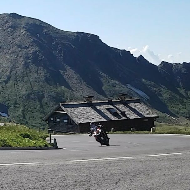 KurvenScout Ein Motorrad fährt durch eine Kehre auf einer sonnigen Straße mit Grünflächen und den Alpenhauptkamm im Hintergrund.